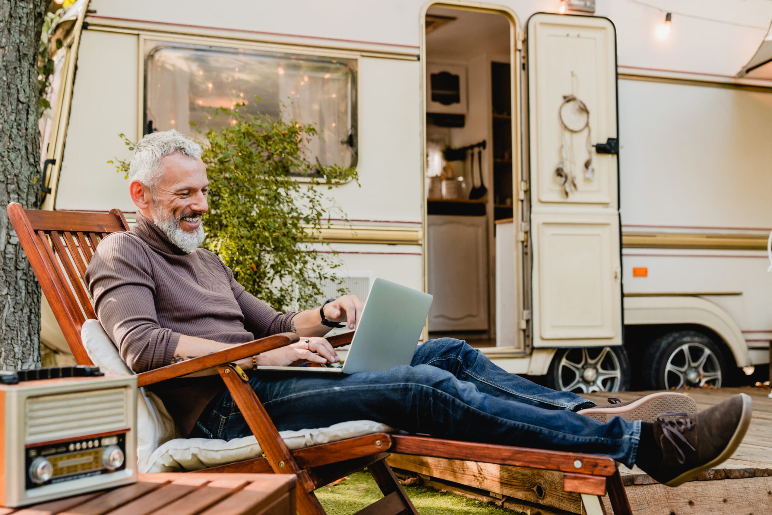 Attractive grey-haired man resting on the wooden deck chair using laptop with caravan van behind